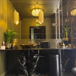 Ładny Dom - Luksusowa łazienka w ciemnej tonacji z marmurowymi elementami, złotą umywalką i dekoracyjnym oświetleniem, widoczne rośliny i złota statuetka
