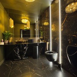 Ładny Dom - Luksusowa łazienka w ciemnej kolorystyce z marmurowymi płytkami, podświetleniem LED i złotymi akcentami, w tym ozdobnymi lampami i umywalką.