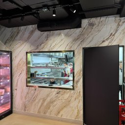 Ładny Dom - Ściana wykończona płytami imitującymi marmur z złotymi żyłami, widok na kuchnię przez okno w ścianie, po lewej chłodziarka z produktami spożywczymi, czarne elementy sufitu i drzwi.