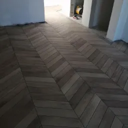 Ułożona w jodełkę, brązowa podłoga z płytek imitujących drewno, w trakcie prac wykończeniowych, widoczny agregat malarski w tle.