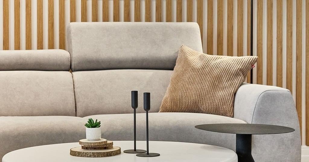 Jasnoszara sofa z regulowanym zagłówkiem, beżowa poduszka z grubym splotem, czarne świeczniki na okrągłym białym stoliku kawowym, mała roślina doniczkowa i dekoracyjna drewniana ścianka z pionowych...