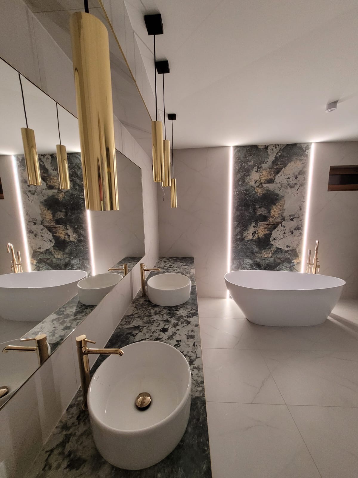 Nowoczesna łazienka z dwiema umywalkami nablatowymi o owalnym kształcie i złotymi bateriami, marmurowym blatem, wolnostojącą wanną, podświetlaną ścianą z motywem kamienia i złotymi lampami wiszącymi.