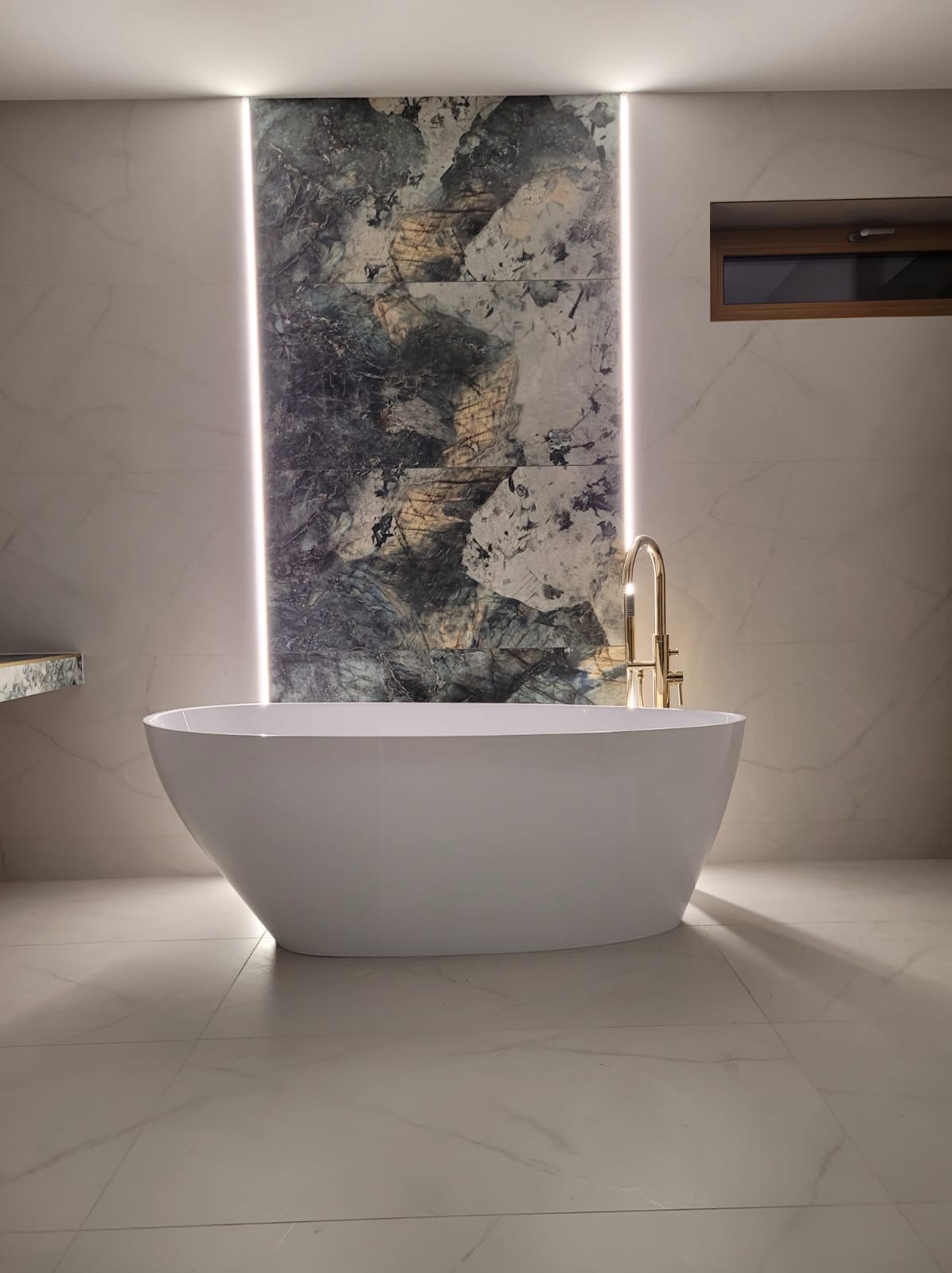 Nowoczesna łazienka z wolnostojącą białą wanną, złotą armaturą i podświetlaną ścianą z płytek o wzorze przypominającym marmur.