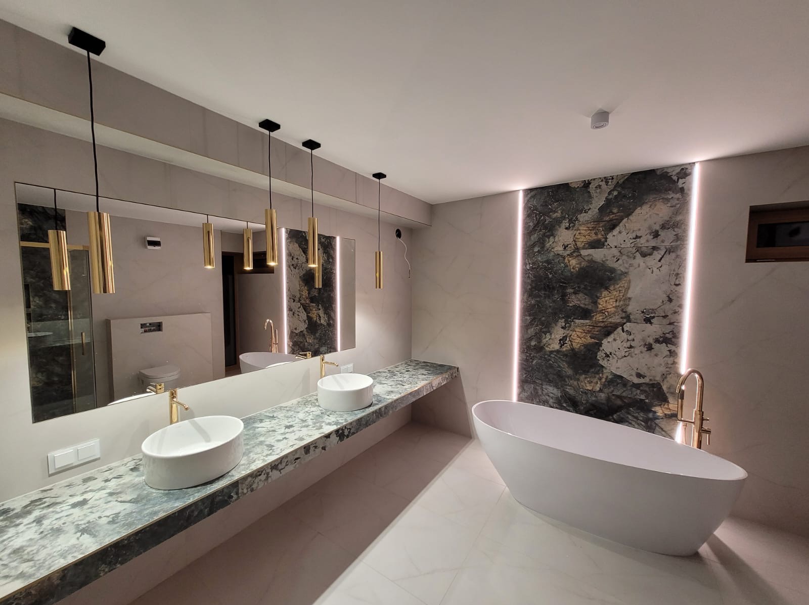 Nowoczesna łazienka z dwoma umywalkami na blacie z szarego marmuru, wolnostojąca wanna z złotą armaturą i podświetlaną ścianą z wzorem kamienia.