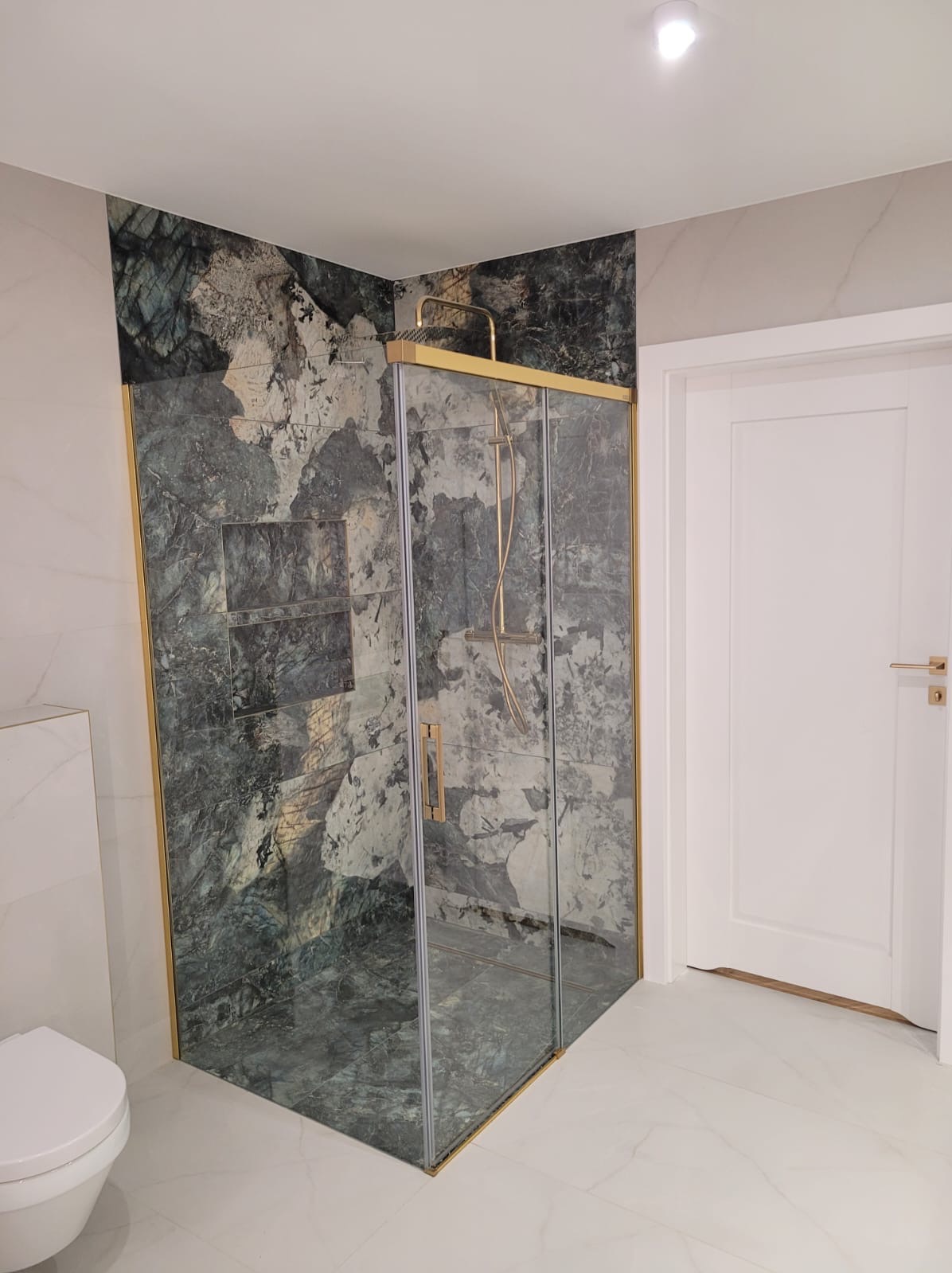 Nowoczesna kabina prysznicowa z grafitową okładziną imitującą kamień, wykończona złotymi profilami, wkomponowana w jasną łazienkę z białą armaturą i drzwiami.