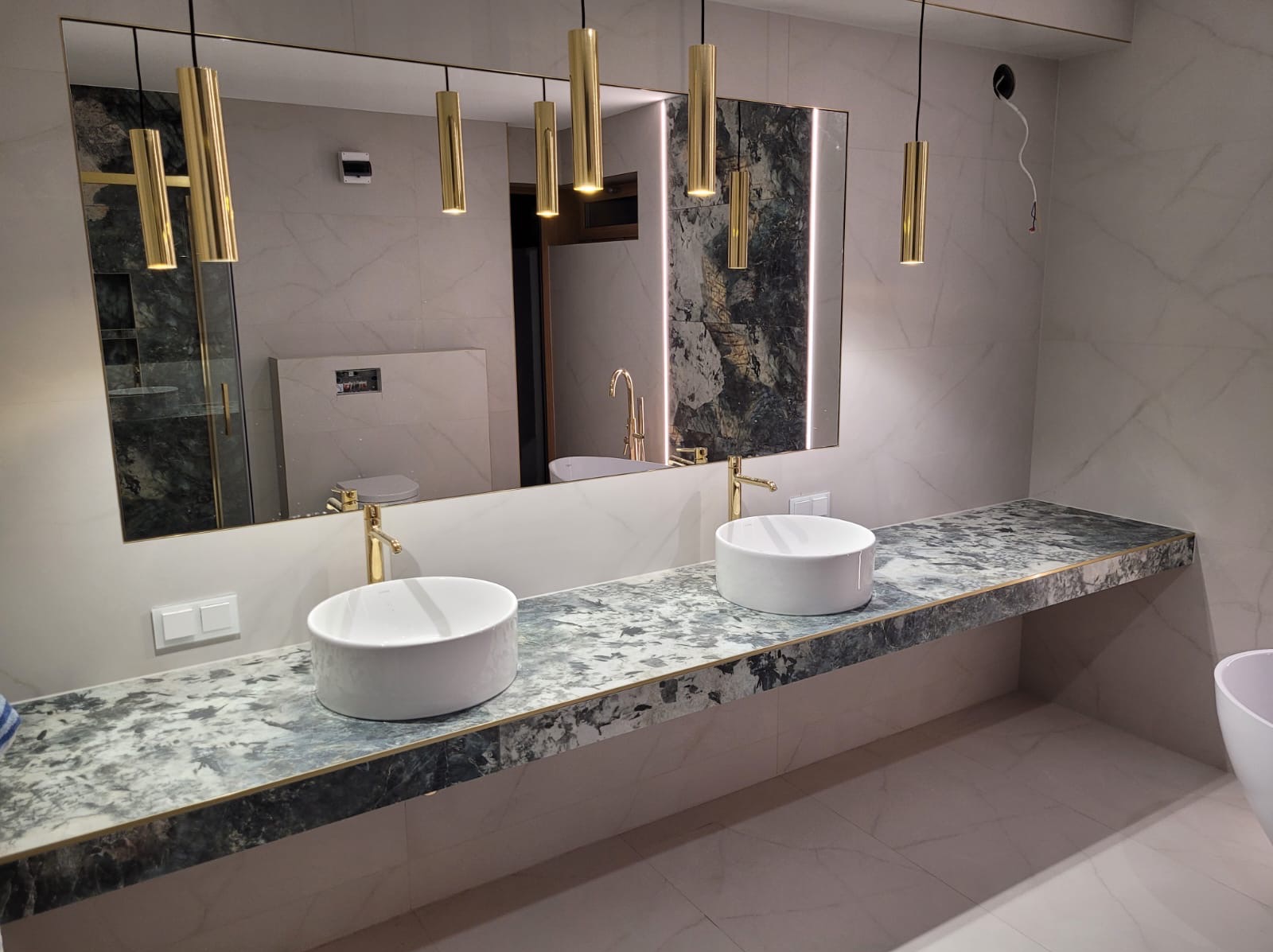 Nowoczesna łazienka z dwoma białymi umywalkami nablatowymi na blacie z szarego marmuru, złotymi bateriami i podwieszanymi lampami w kształcie złotych tub, duże lustro z podświetleniem i widoczną...