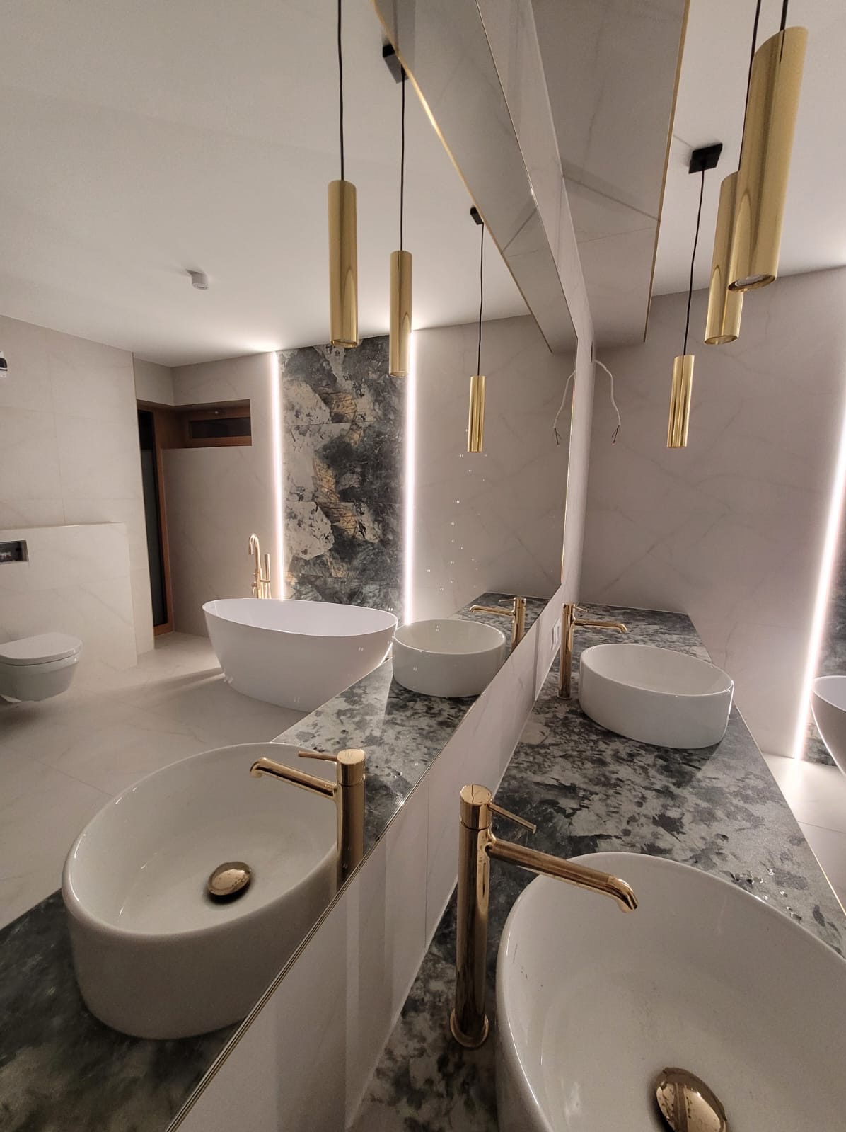 Nowoczesna łazienka z owalnymi umywalkami na marmurowym blacie, złotymi bateriami i wiszącymi lampami w kształcie złotych tub, podświetlona ściana z efektem kamienia.