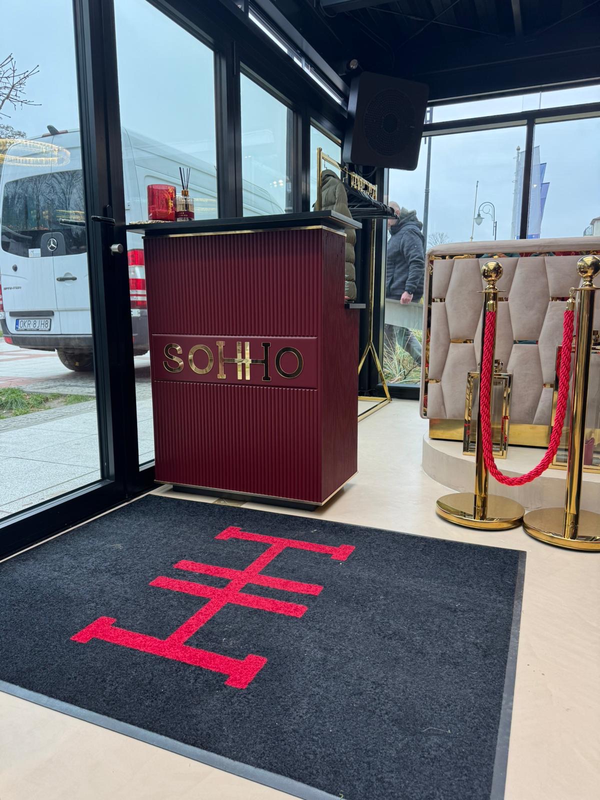 Recepcja z bordowym panelem z napisem SOHO w złotych literach, czarny dywanik z czerwonym logo, złote słupki z czerwonym sznurem, widok na ulicę przez okno, biały samochód dostawczy zaparkowany...