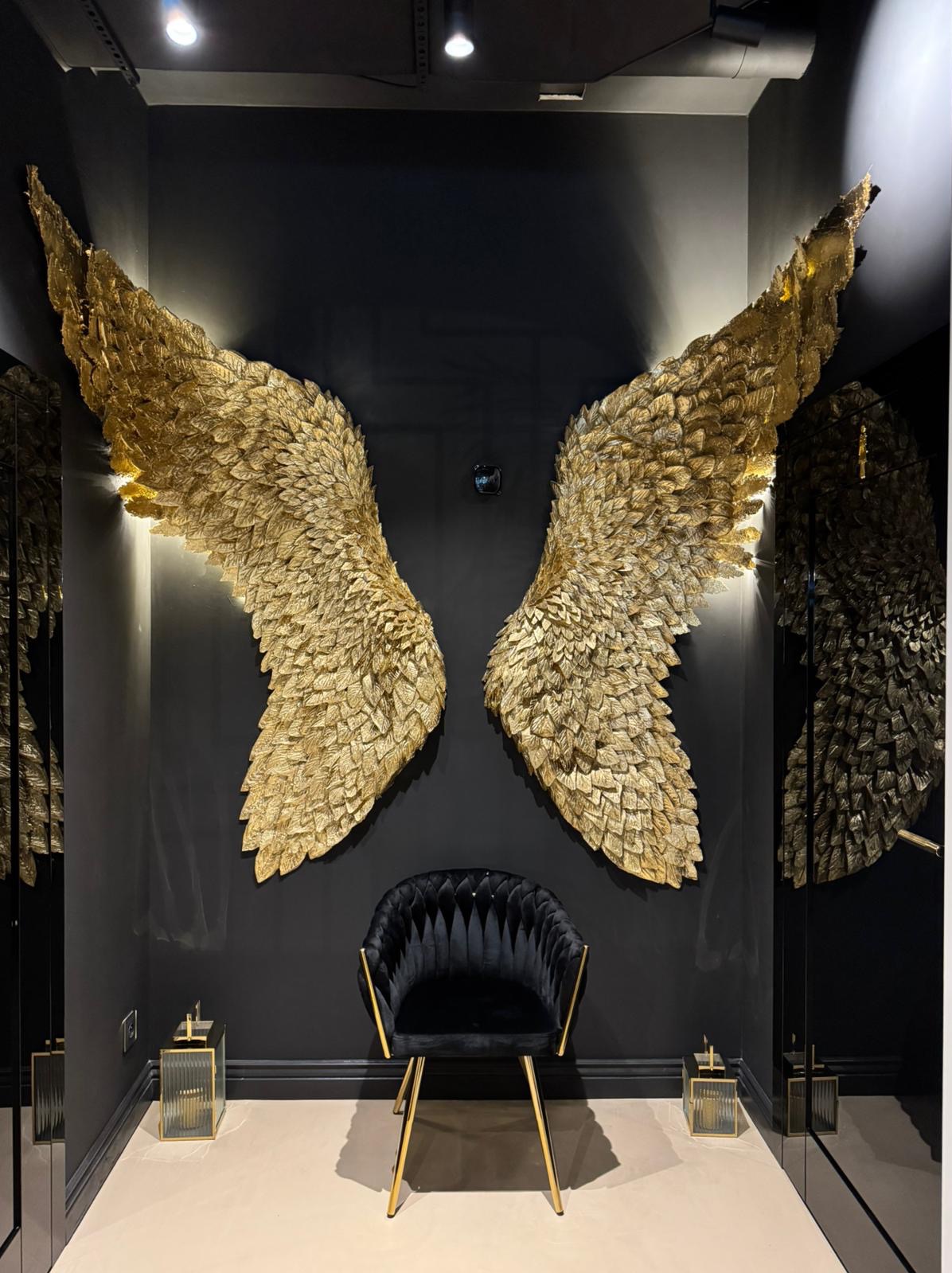 Dekoracyjne złote skrzydła anioła na czarnej ścianie nad czarnym, welurowym krzesłem ze złotymi nogami, oświetlone lampkami, tworzące elegancką aranżację wnętrza.