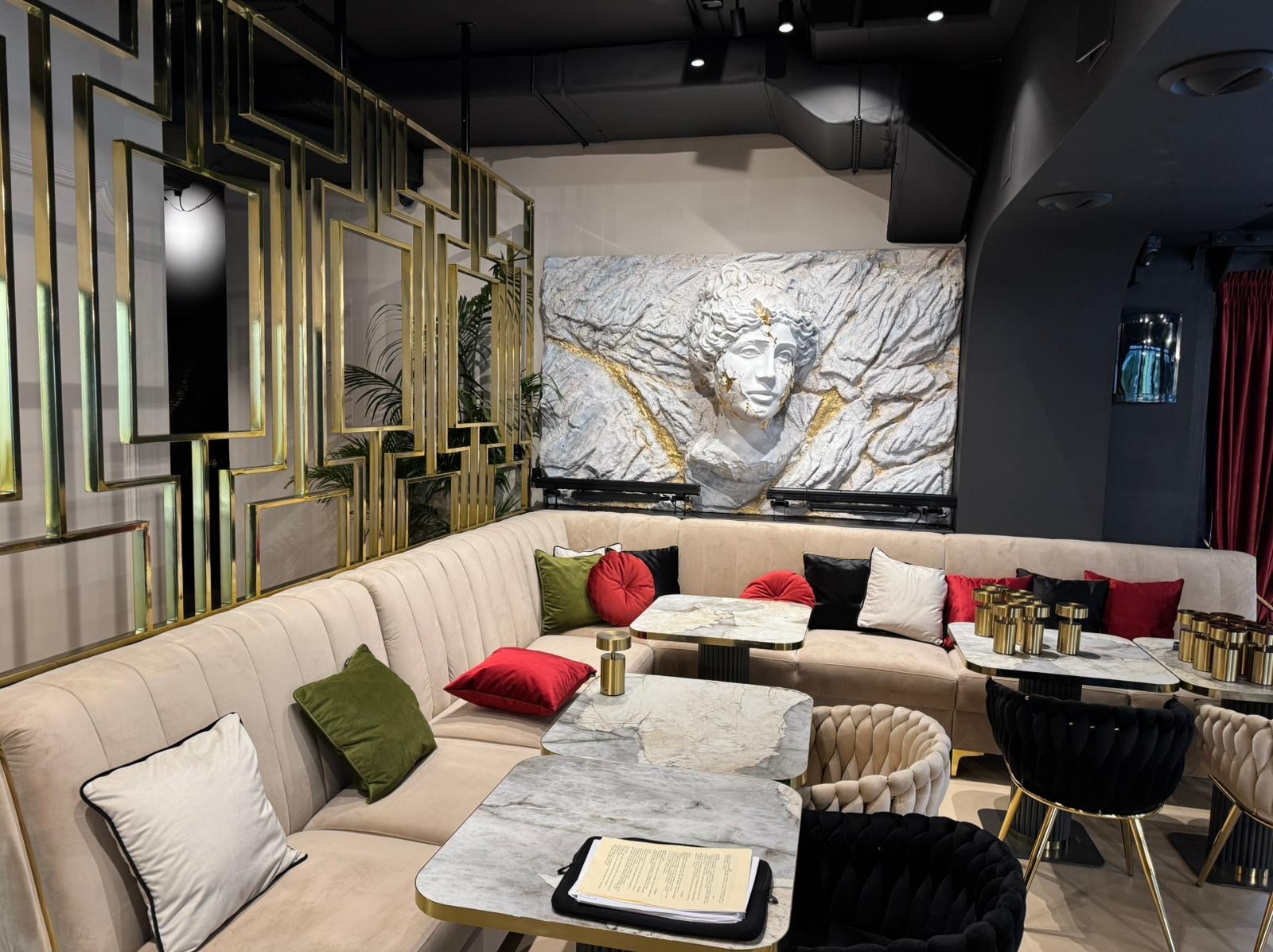 Eleganckie wykończenie wnętrza restauracji z beżową sofą narożną, marmurowymi stolikami i dekoracyjną płaskorzeźbą głowy w stylu antycznym na ścianie.