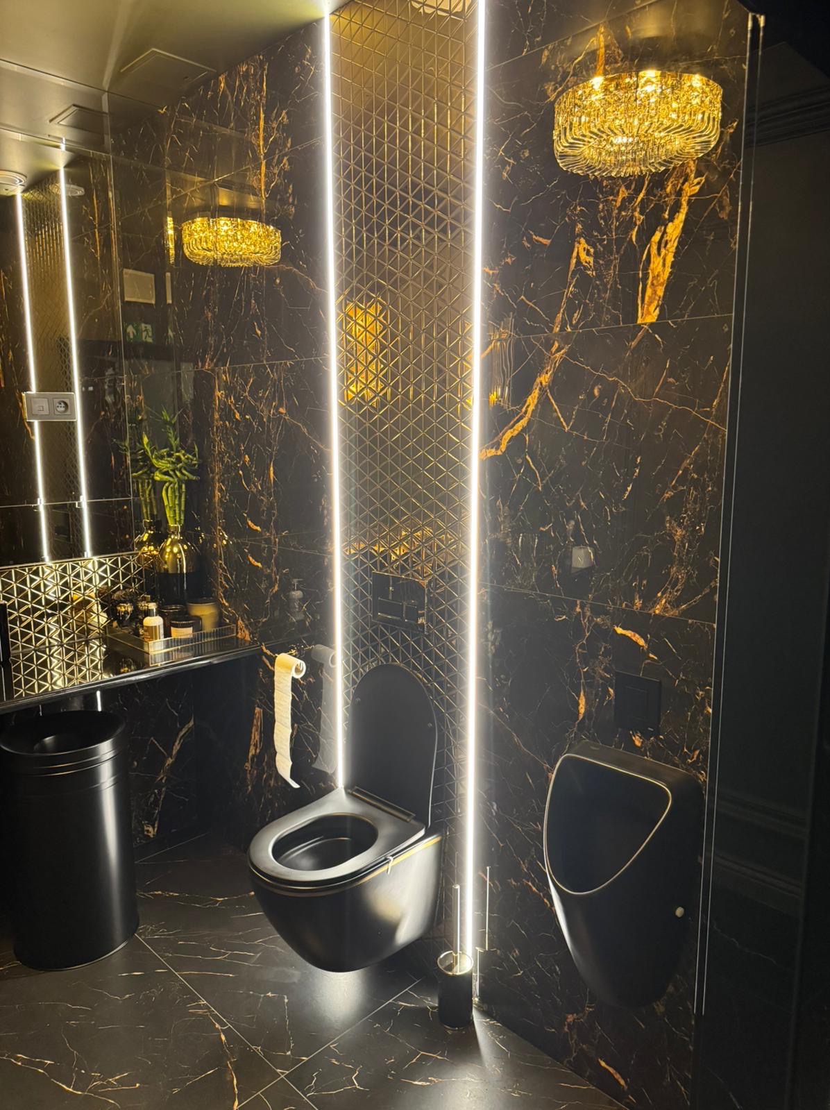 Elegancka, czarna łazienka z marmurowymi płytkami, podświetleniem LED, wiszącą toaletą i pisuar, oświetlona ozdobnym żyrandolem.