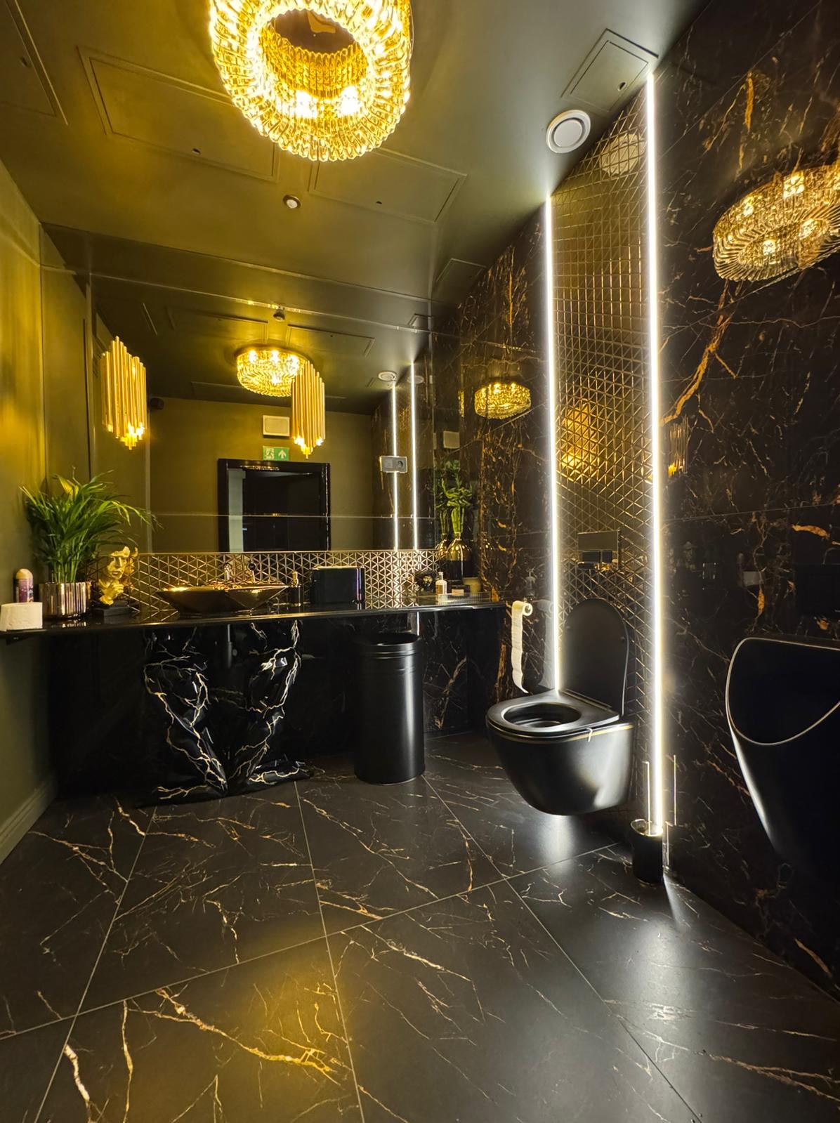 Luksusowa łazienka w ciemnej kolorystyce z marmurowymi płytkami, podświetleniem LED i złotymi akcentami, w tym ozdobnymi lampami i umywalką.