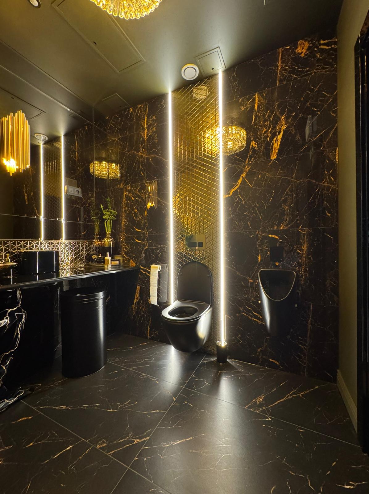 Luksusowa toaleta w czerni i złocie: marmurowe płytki ścienne i podłogowe z wyraźnym żyłowaniem, podświetlane pionowe panele, wisząca muszla klozetowa i pisuar w tym samym kolorze, kryształowe...