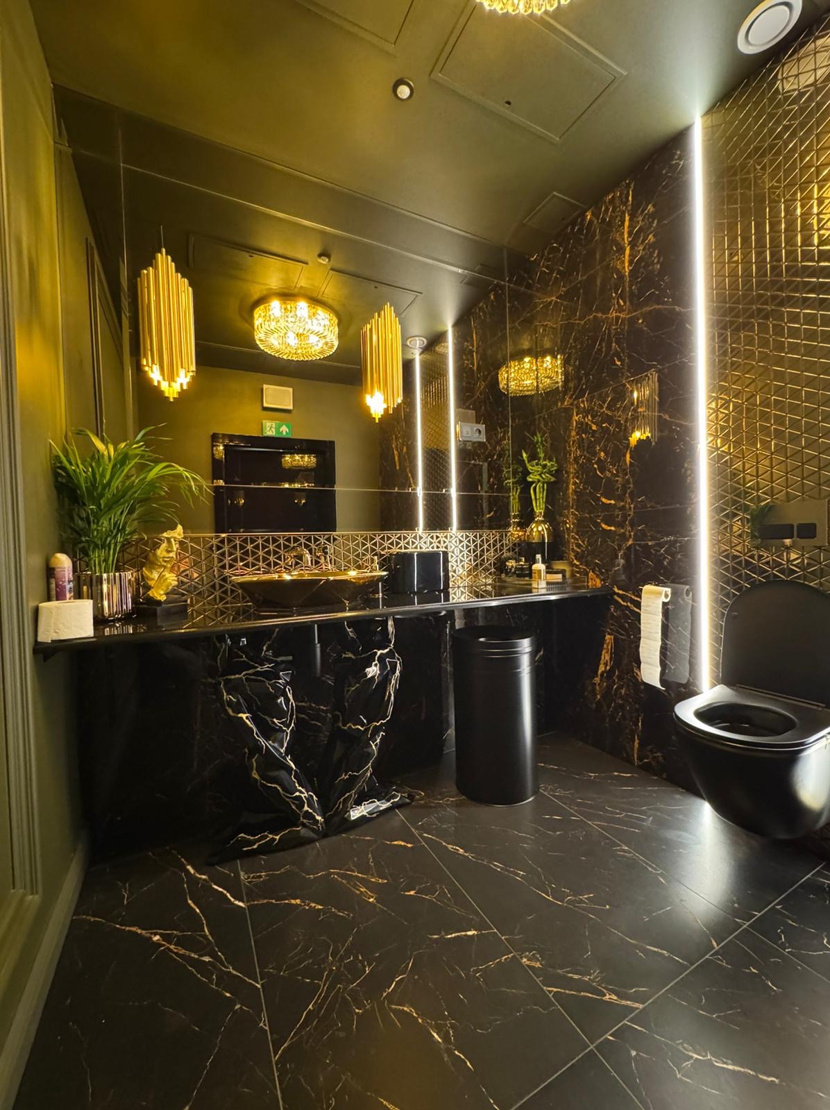 Luksusowa łazienka z czarnym marmurem na ścianach i podłodze, złotymi akcentami w oświetleniu i armaturze, oraz toaletą w kolorze czarnym.