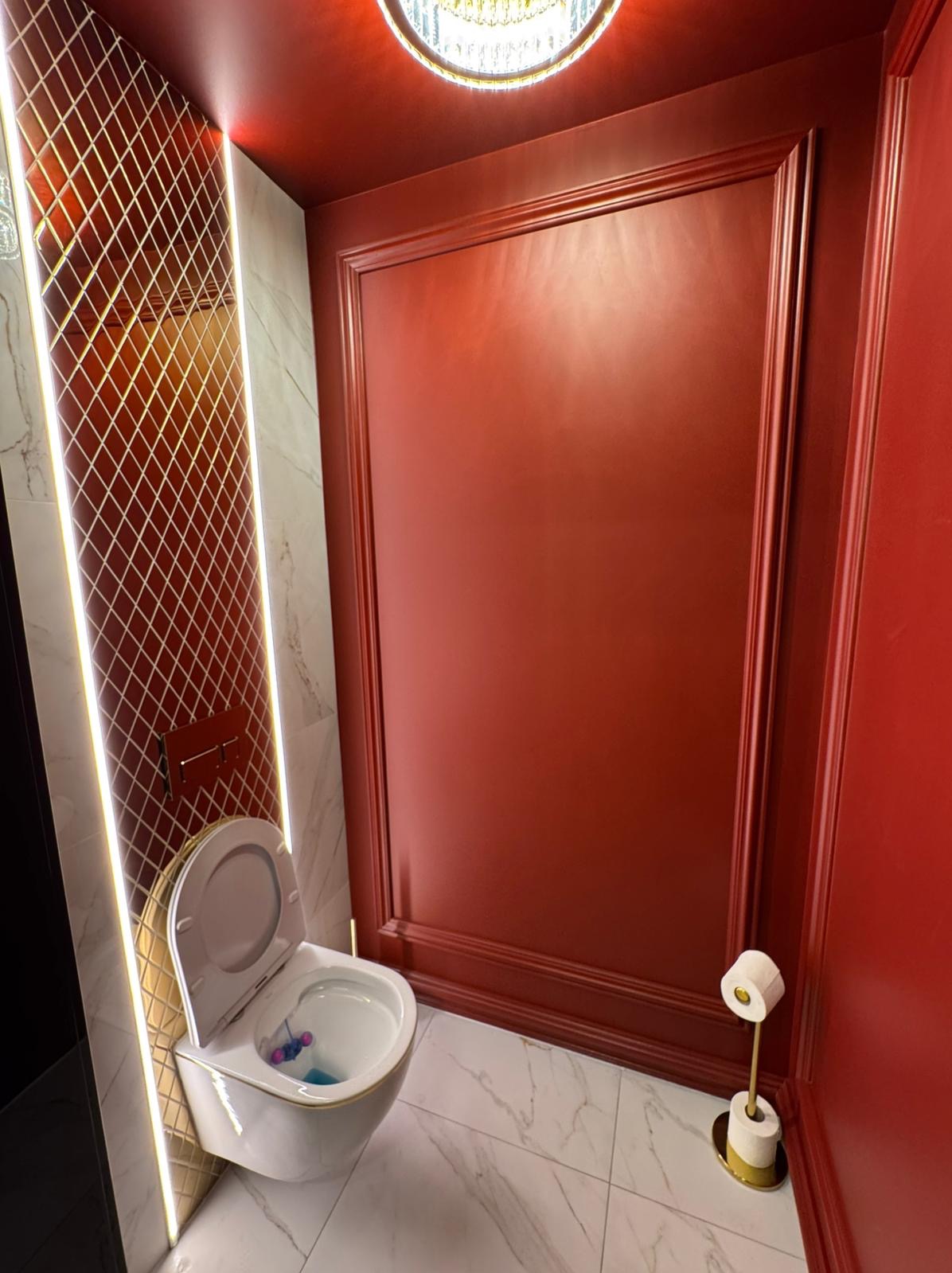 Wnętrze łazienki w odcieniach czerwieni i złota, z marmurową podłogą, toaleta wisząca z otwartą klapą i kostką do WC, ściana z czerwonymi panelami i oświetlona ściana z mozaiki w romby.