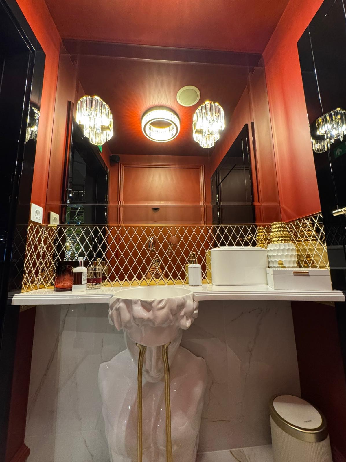 Ekskluzywna łazienka w odcieniach czerwieni, z umywalką w kształcie popiersia, złotymi akcentami na mozaice i dekoracyjnych lampach kryształowych.