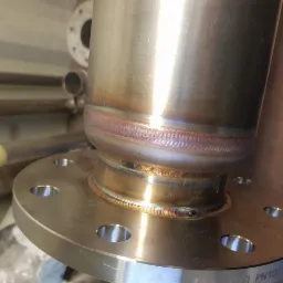 Precyzyjnie wykonane spawy na złączu kołnierzowym rury ze stali nierdzewnej, widoczne ślady termiczne po spawaniu, z rurami w tle.