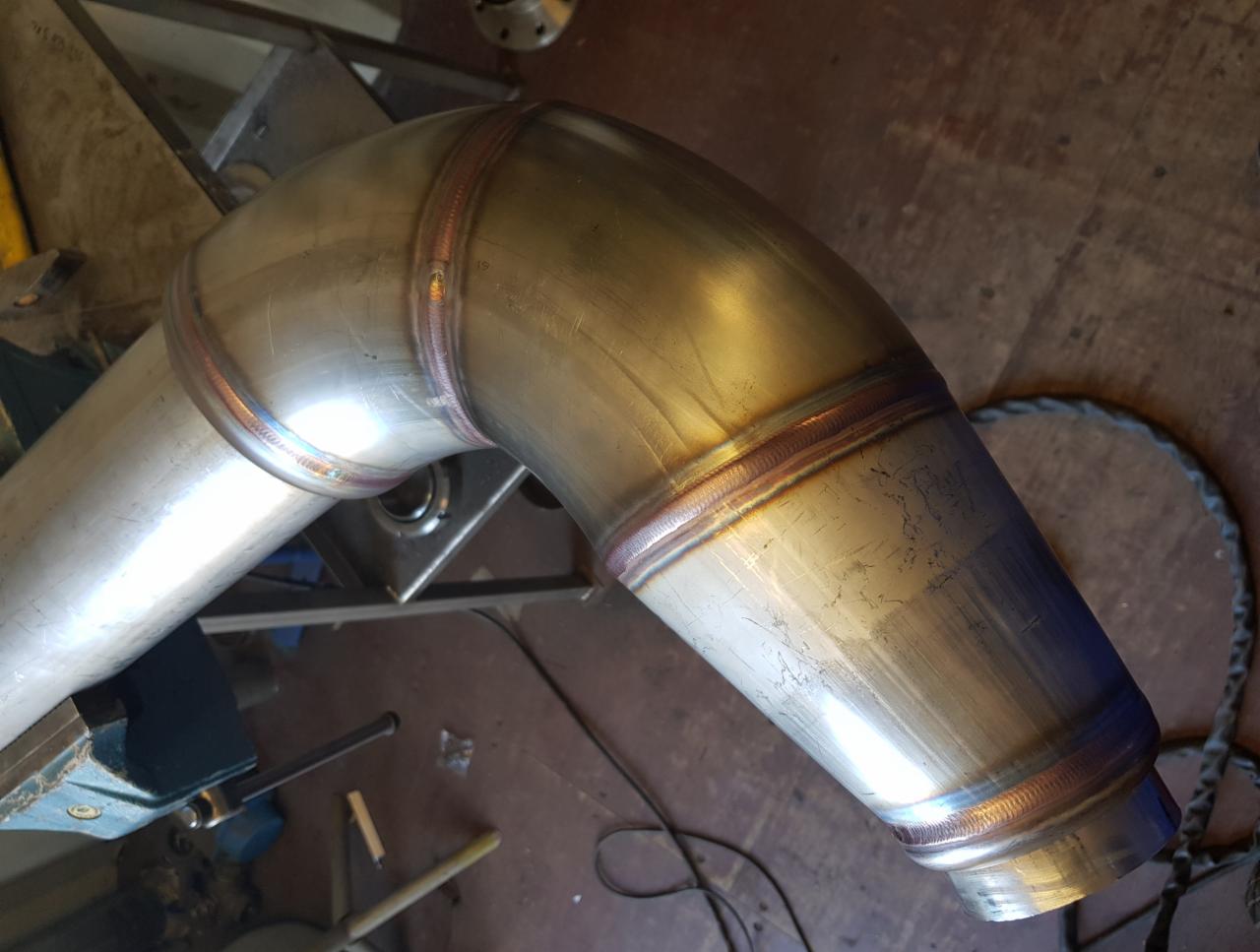 Spawany kolanko z rury nierdzewnej, widoczne spoiny i opalizujące przebarwienia termiczne, zamocowane w imadle.