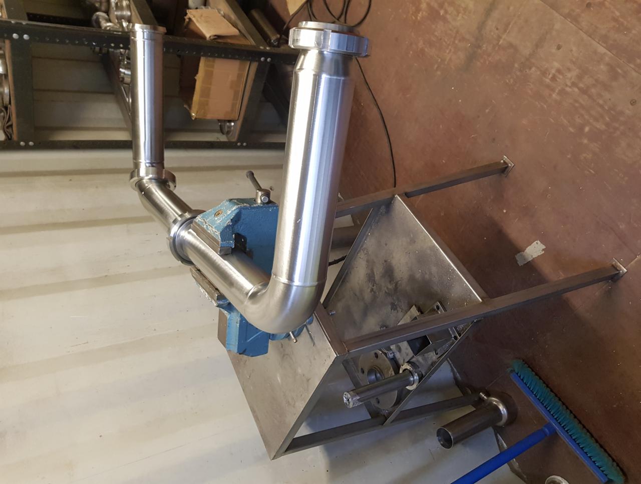 Konstrukcja ze stali nierdzewnej, rury gięte i spawane, zamocowane w imadle ślusarskim, na metalowym stole warsztatowym, w tle regał z kartonami.