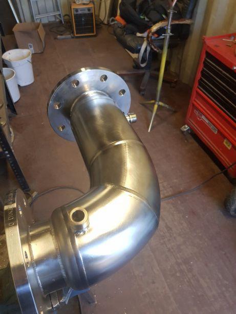 Błyszczący, spawany kolanko rury ze stali nierdzewnej z kołnierzami, leżące na podłodze warsztatu, widoczny czerwony wózek narzędziowy w tle.