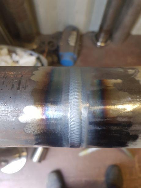 Metalowa rura z widocznym świeżym spoiwem spawalniczym, tęczowe przebarwienia na metalu w pobliżu złącza, w tle narzędzia i fragment obuwia.