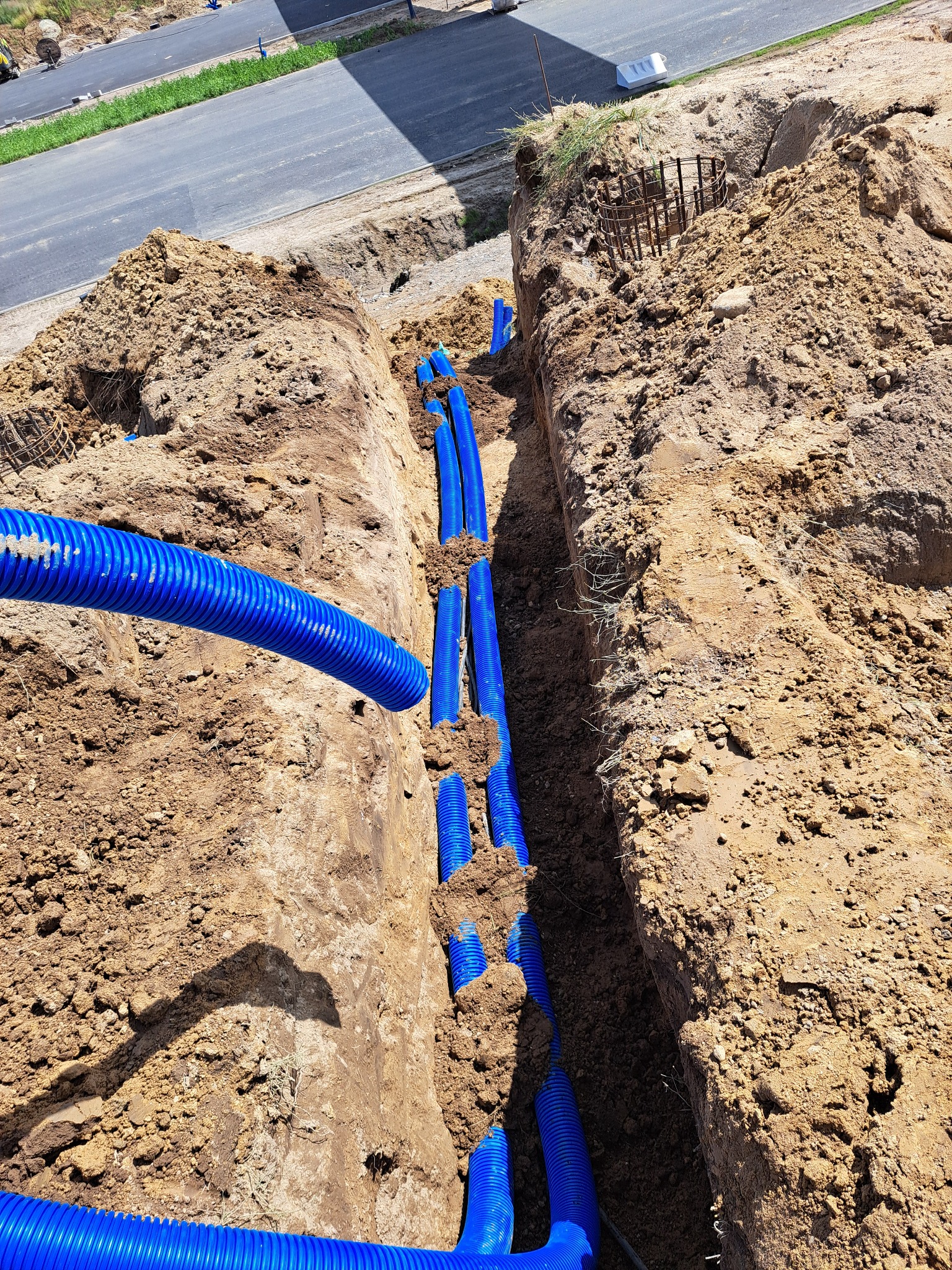 Wąski wykop z niebieskimi rurami karbowanymi ułożonymi wzdłuż, ziemia po bokach, fragment drogi w tle. Widoczne zbrojenie w ziemi.