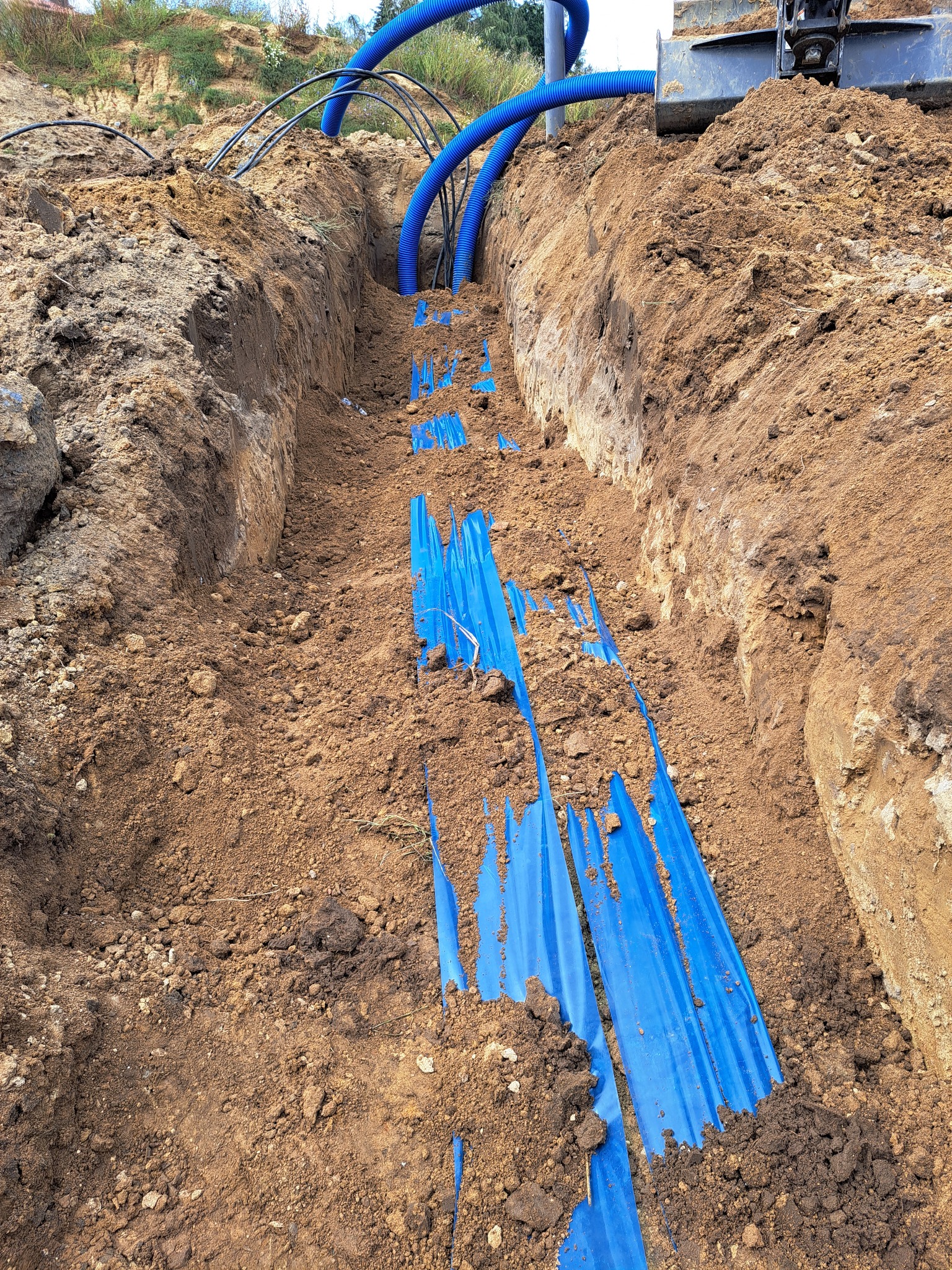 Wykopany rów z niebieskimi rurami karbowanymi na dnie, częściowo przykrytymi folią. Na górze widoczna krawędź wykopu i fragment koparki.