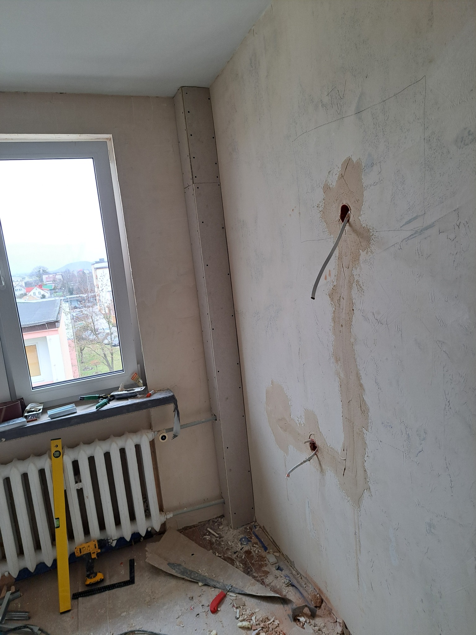 Wnętrze w trakcie remontu: ściana z instalacją elektryczną, częściowo położona gładź, przy oknie grzejnik, poziomica i narzędzia. Widoczne płyty karton-gips.