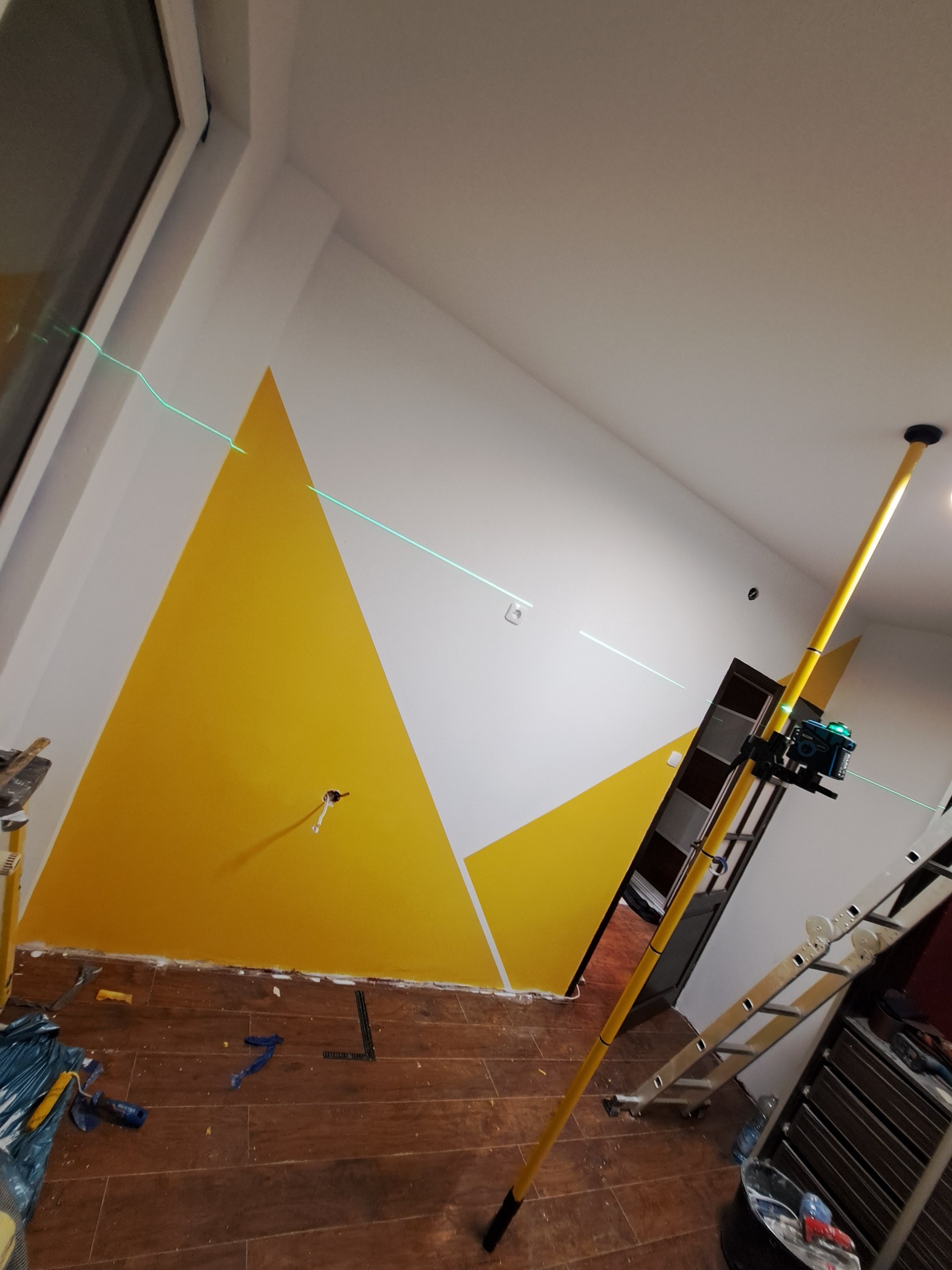 Ściana po malowaniu z geometrycznym wzorem w odcieniach żółci, wyznaczonym precyzyjnie za pomocą lasera. Widoczne narzędzia malarskie i drabina. Podłoga wykończona panelami.