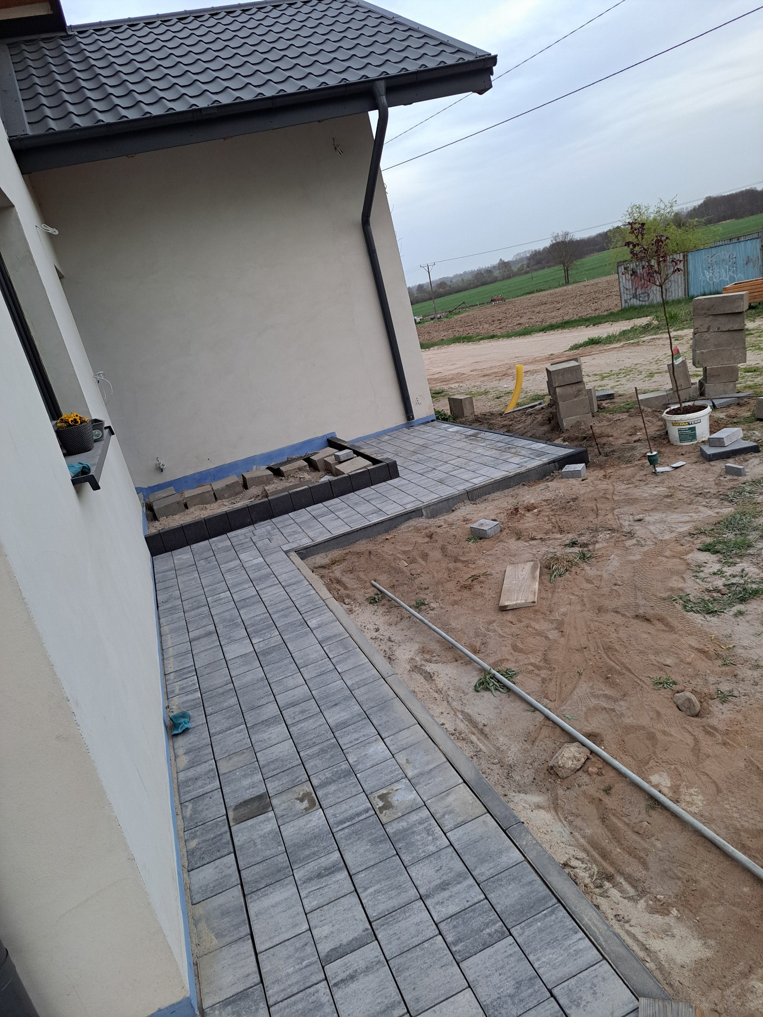 Układanie szarej kostki brukowej przy ścianie budynku, widoczny fragment ułożonej nawierzchni i etap prac z podsypką. W tle pole i budynki gospodarcze.