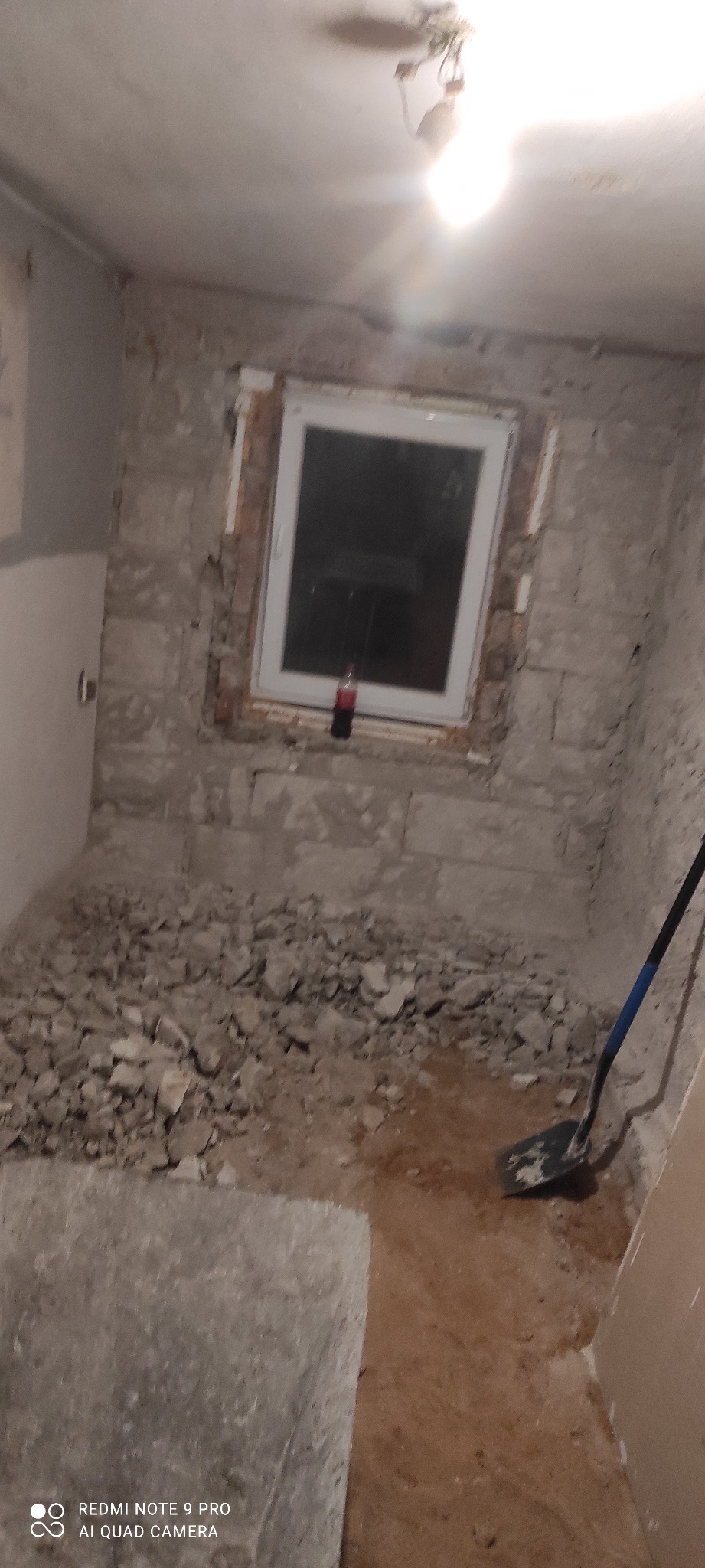 Wnętrze w trakcie remontu: ściany z odsłoniętą cegłą, gruz na podłodze, widoczna łopata. Okno z butelką. Oświetlenie sufitowe.