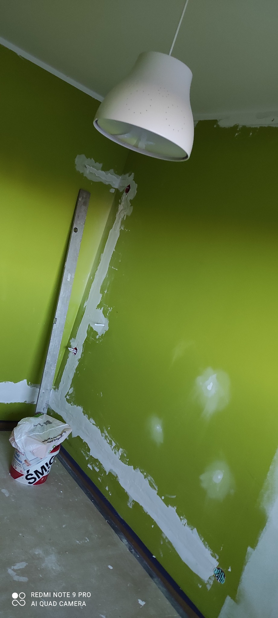 Zielona ściana w trakcie szpachlowania, widoczny poziomica i wiadro z masą szpachlową. W tle biała lampa zwisająca z sufitu.