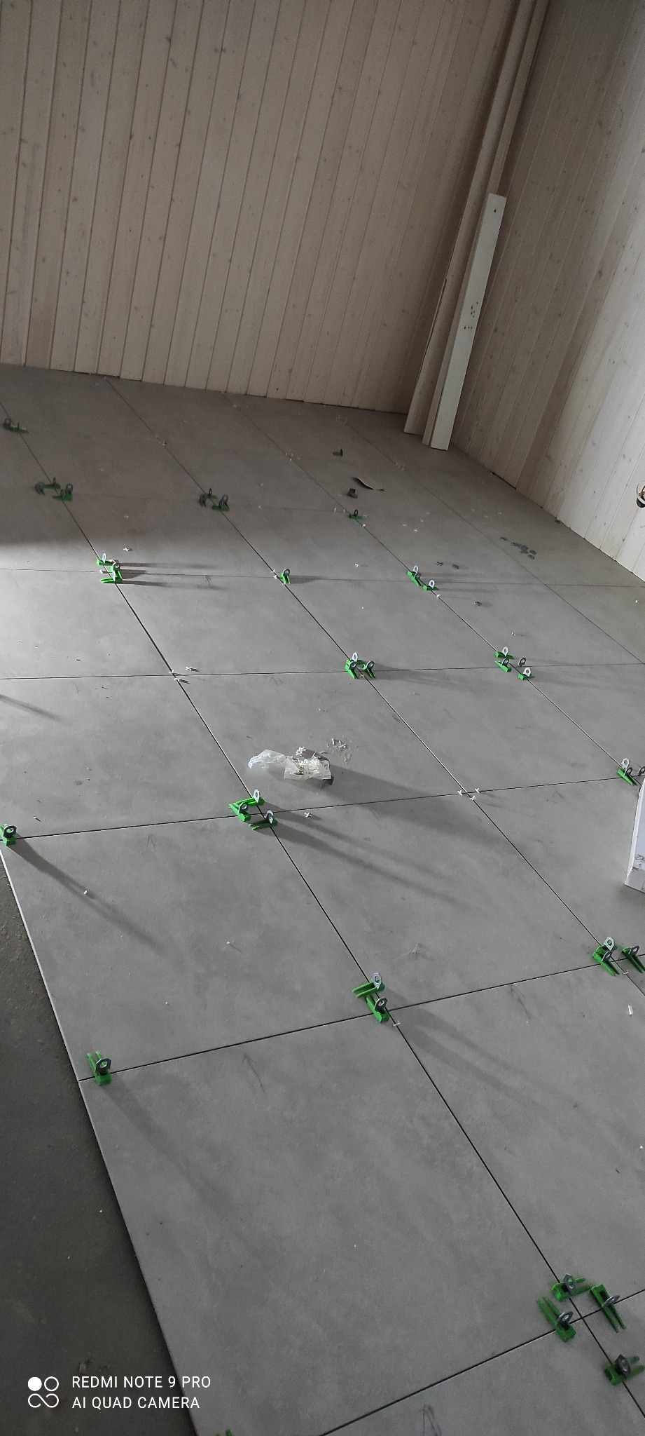 Układanie szarych płytek podłogowych z użyciem zielonych klipsów poziomujących. Widoczne drewniane panele ścienne. Etap prac wykończeniowych, wnętrze w trakcie realizacji.