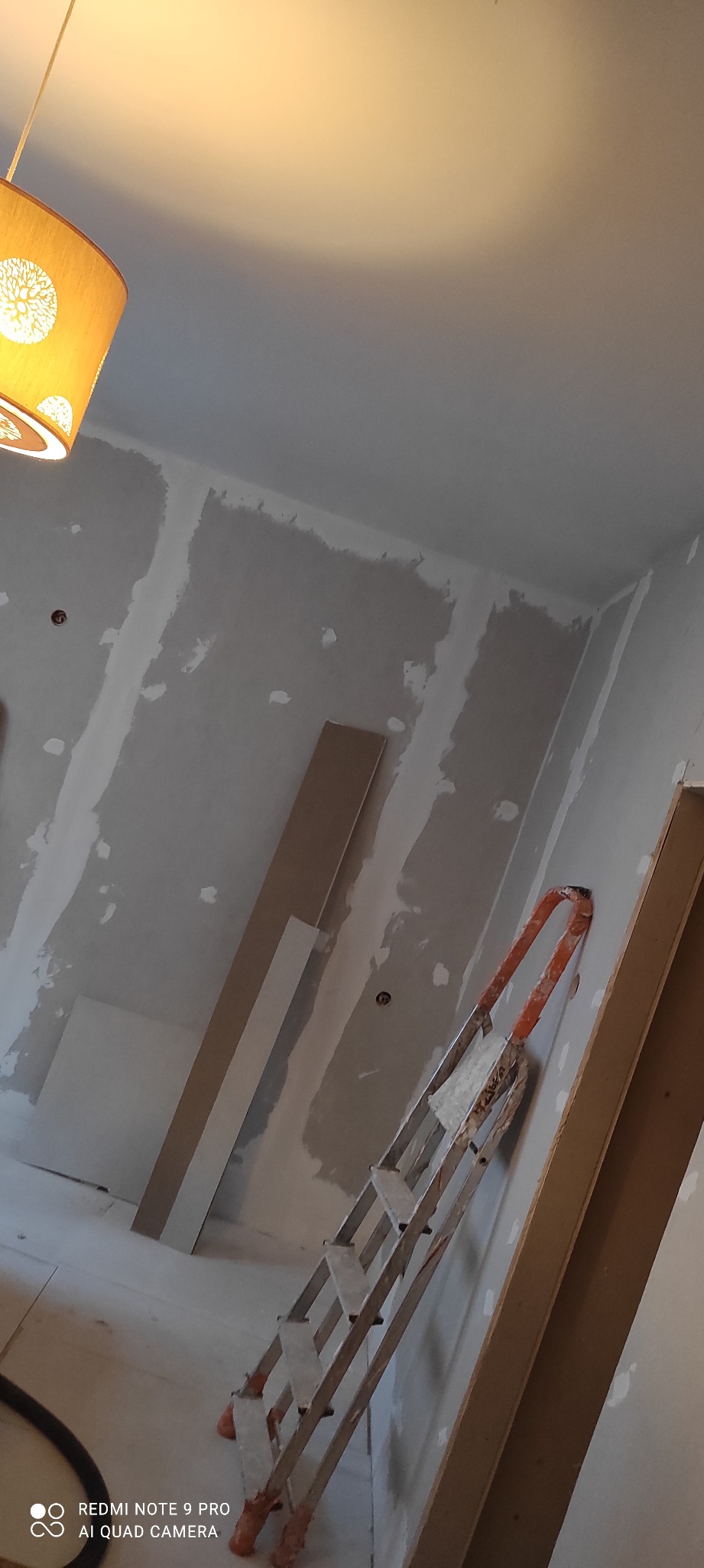 Pokój w trakcie remontu: ściany z widocznymi łączeniami płyt gipsowo-kartonowych, drabina oparta o ścianę, widoczne profile i lampa. Ujęcie z dołu, ciepłe oświetlenie.