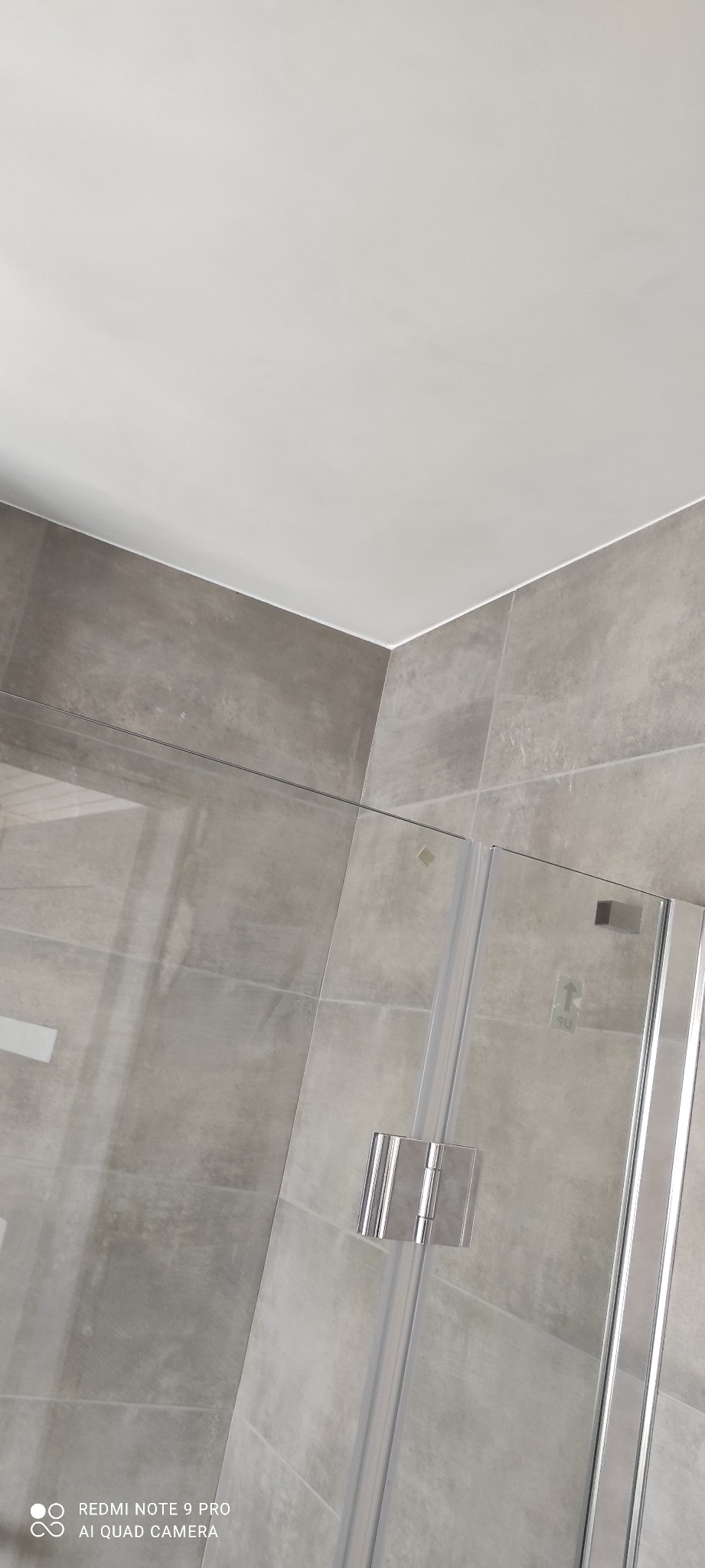 Szklana kabina prysznicowa z chromowanymi zawiasami na tle szarych, kwadratowych płytek. Minimalistyczny design, jasne fugi, biały sufit. Ujęcie z dołu.