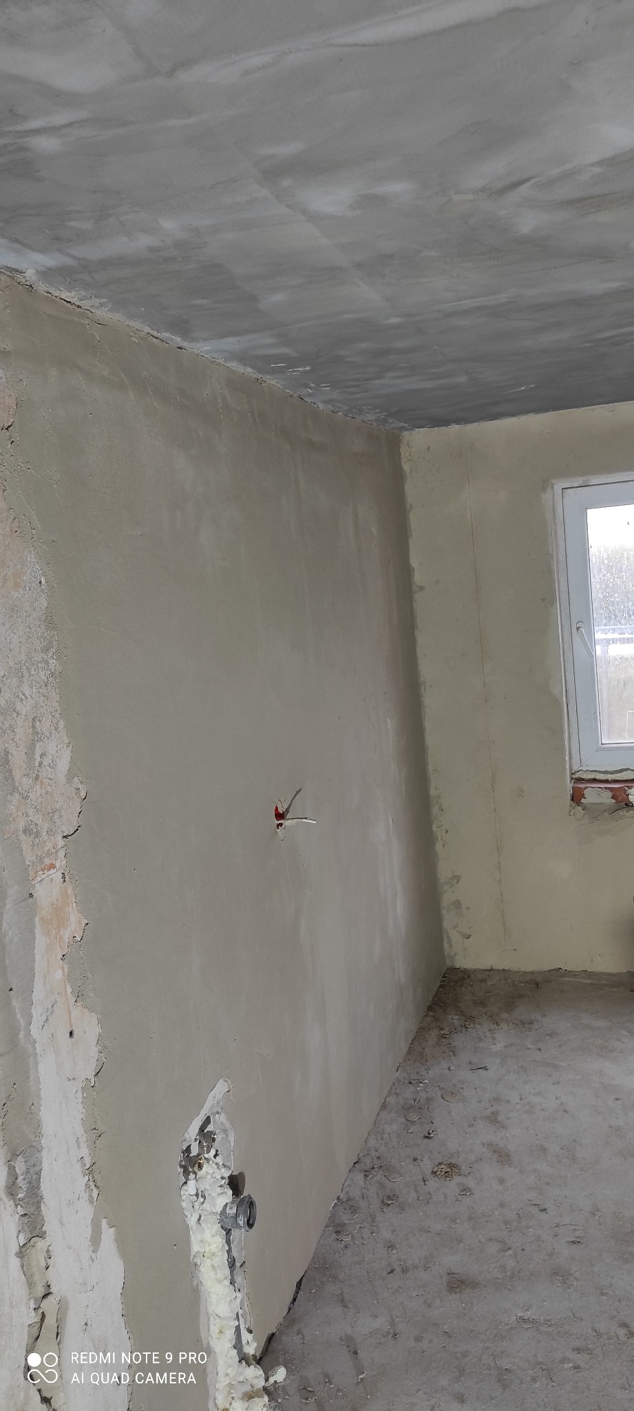 Surowy pokój po szpachlowaniu: ściany w szarej gładzi, widoczne instalacje elektryczne i hydrauliczne. Okno z białymi ramami w tle. Podłoga betonowa.