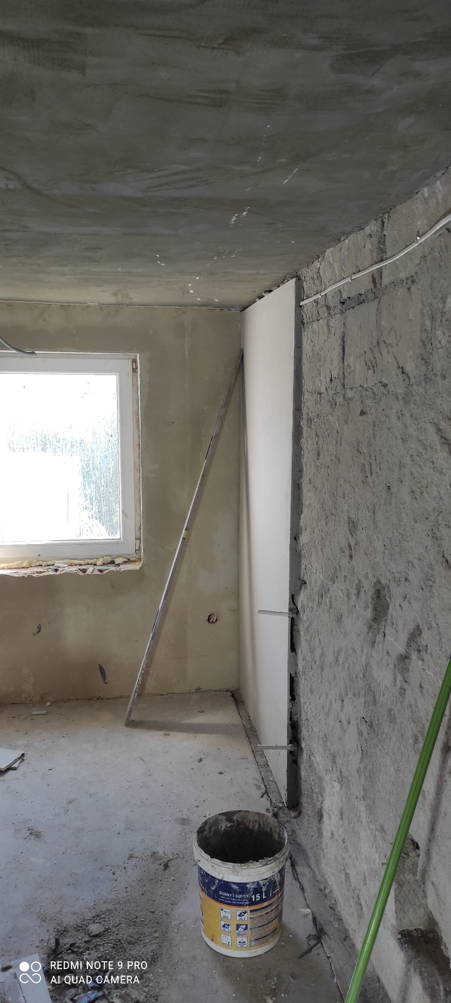 Montaż płyty G-K na nierównej ścianie w surowym wnętrzu. Widoczny poziomica i wiadro z farbą, co sugeruje etap prac wykończeniowych. Surowy stan ścian i sufitu.