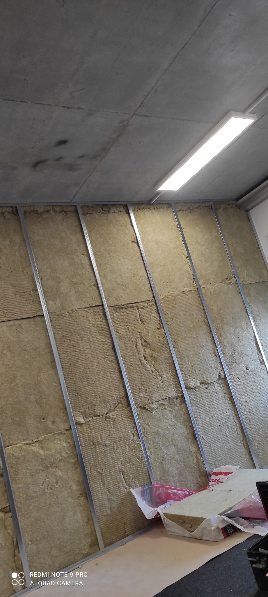 Montaż izolacji z wełny mineralnej w metalowym stelażu przy suficie. Widoczne pakiety wełny. Ujęcie z dołu, lekko pod kątem.