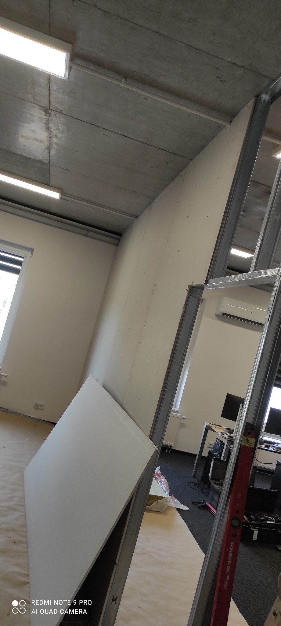 Montaż ścianki działowej z płyty gipsowo-kartonowej na metalowym stelażu w biurze. Widoczny poziomica, surowy betonowy sufit i okno.
