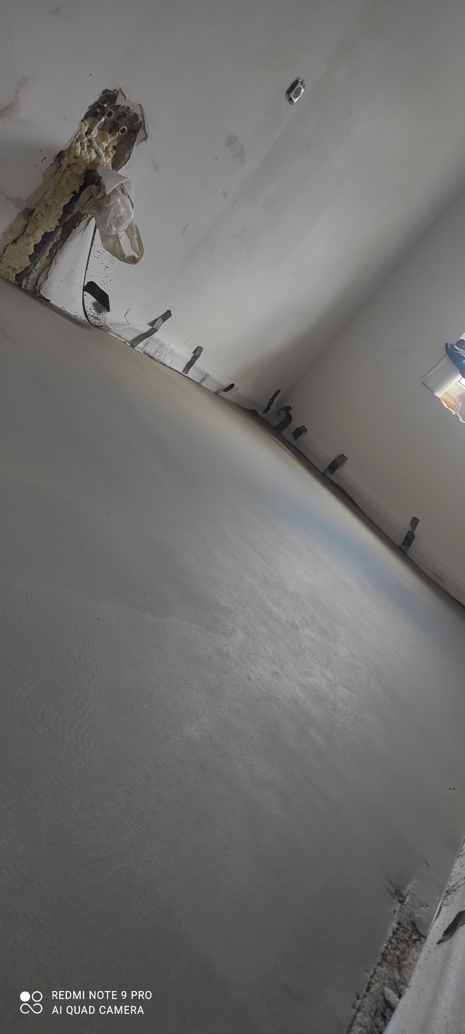 Świeżo nałożona, gładka szpachla na ścianie w trakcie remontu. Widoczne wystające elementy montażowe i pianka uszczelniająca przy suficie. Ujęcie pod kątem.