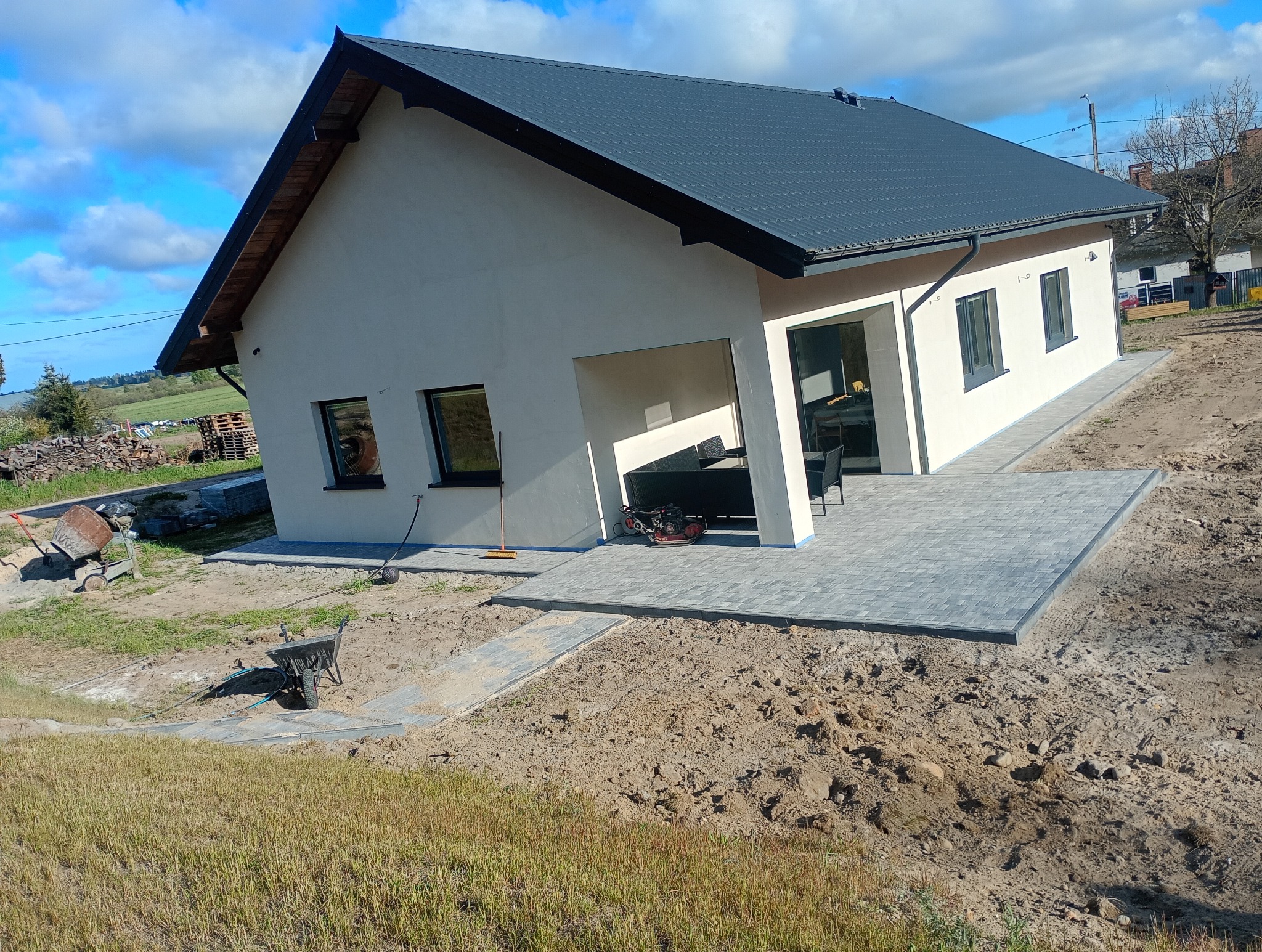 Ułożona kostka brukowa na tarasie i ścieżce w Mrzezinie. Nowoczesny dom jednorodzinny w tle. Prace brukarskie w toku, widoczny sprzęt budowlany.