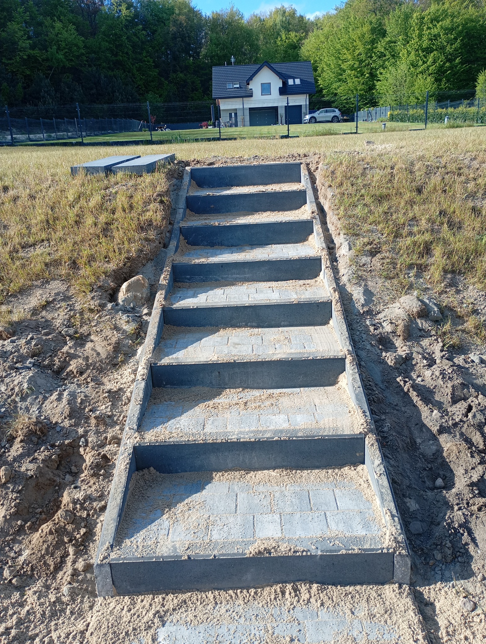 Nowe schody z kostki brukowej prowadzą w górę zbocza, z domem w tle. Widoczne świeże prace ziemne i piasek na stopniach. Układanie kostki brukowej w ogrodzie.
