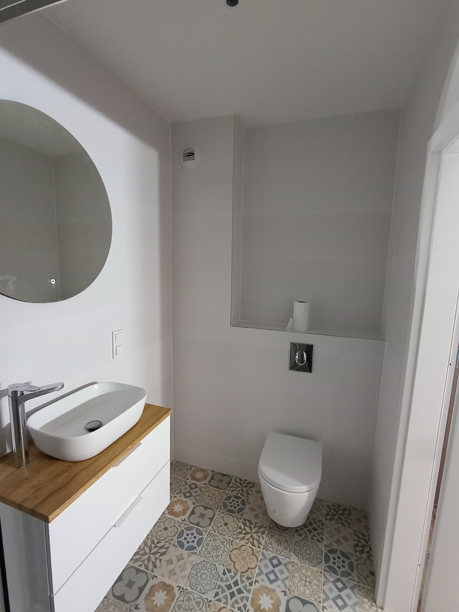 Nowoczesna łazienka w bieli z okrągłym lustrem, umywalką na szafce z drewnianym blatem i wzorzystą podłogą. Minimalistyczny design z ukrytą spłuczką WC.