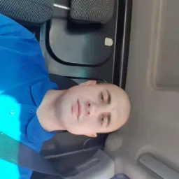 Mężczyzna w niebieskiej koszuli polo siedzący w pojeździe, z widocznym zagłówkiem i pasem bezpieczeństwa.