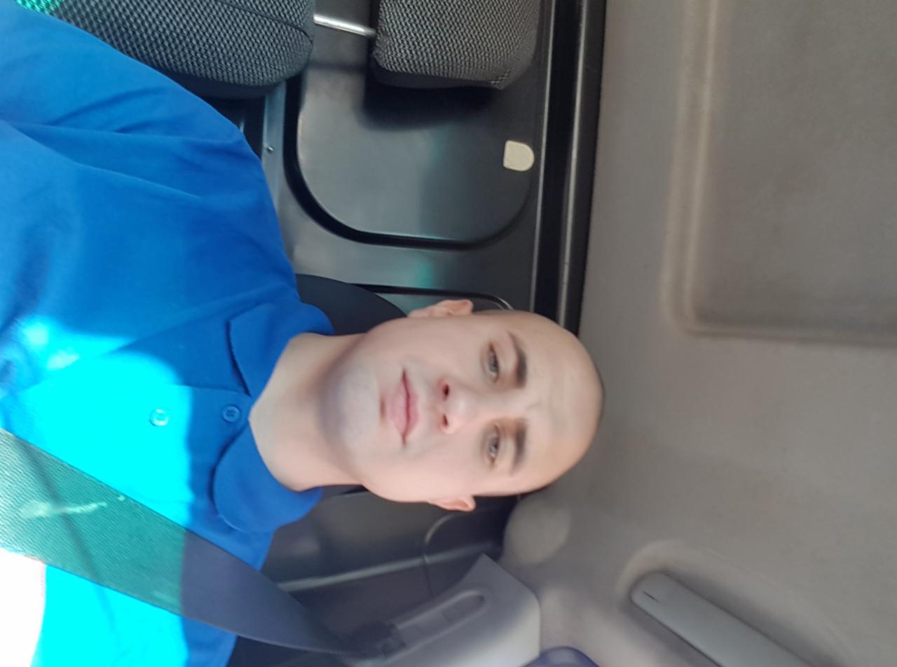 Mężczyzna w niebieskiej koszuli polo siedzący w pojeździe, z widocznym zagłówkiem i pasem bezpieczeństwa.