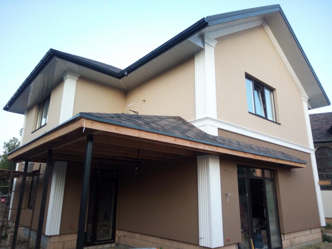 Dwukondygnacyjny dom z beżową elewacją, białymi dekoracyjnymi kolumnami i ciemnym dachem. Widoczny fragment tarasu z drewnianą konstrukcją i czarnymi słupami.