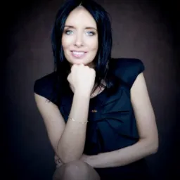 Portret uśmiechniętej kobiety z czarnymi włosami i niebieskimi oczami, opierającej brodę na dłoni, w czarnej sukience z kokardą, na ciemnobrązowym tle.