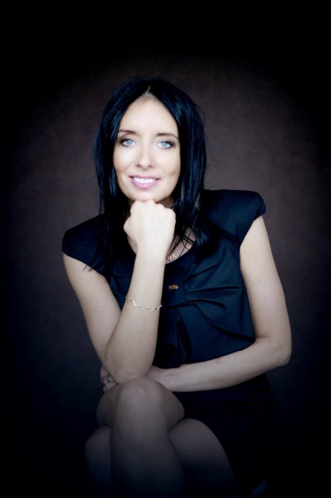 Portret uśmiechniętej kobiety z czarnymi włosami i niebieskimi oczami, opierającej brodę na dłoni, w czarnej sukience z kokardą, na ciemnobrązowym tle.