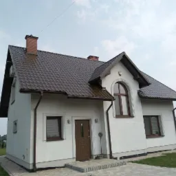 Dom jednorodzinny z brązowym dachem z blachodachówki, białymi ścianami elewacji i brązowymi rynnami, widok z niskiej perspektywy.