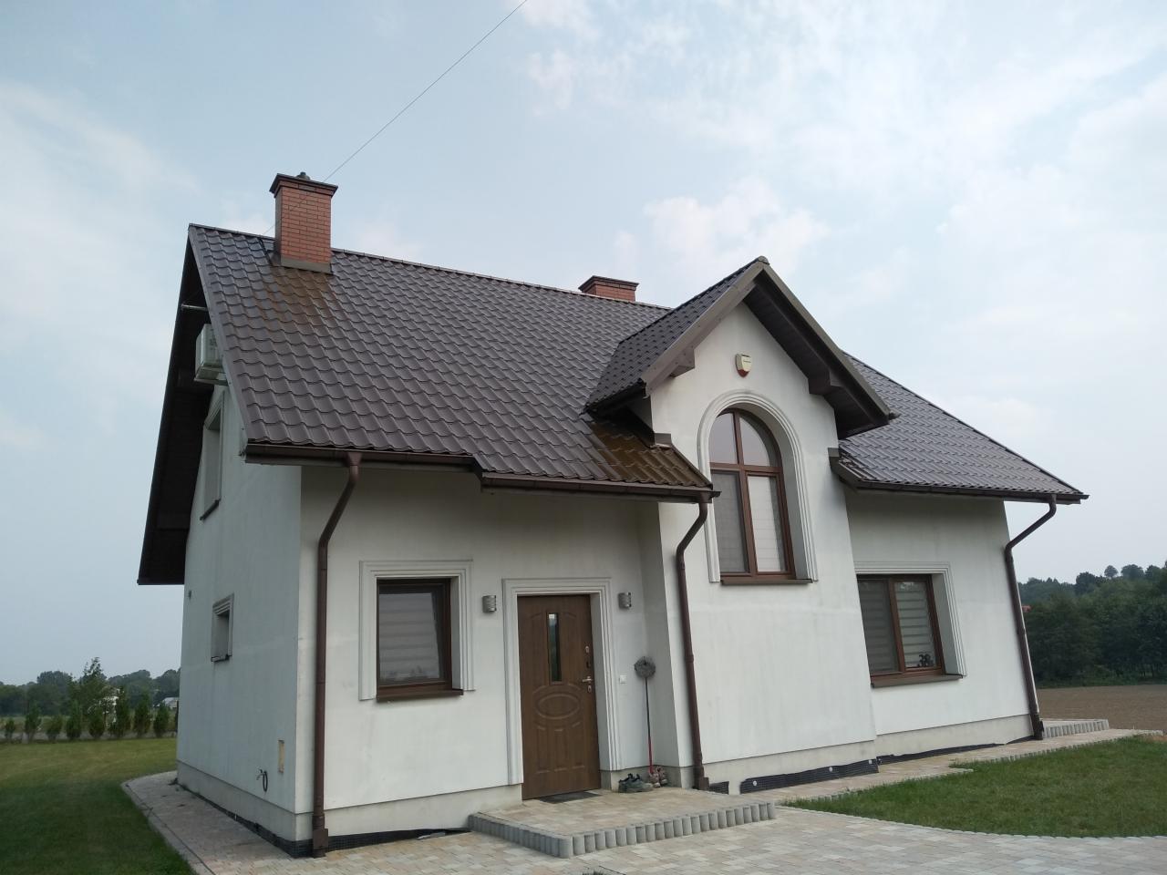 Dom jednorodzinny z brązowym dachem z blachodachówki, białymi ścianami elewacji i brązowymi rynnami, widok z niskiej perspektywy.